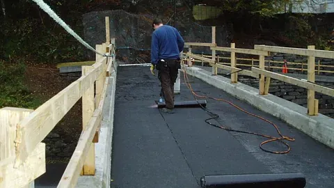 Mann ruller ut asfaltpapp på gangbru med midlertidig rekkverk